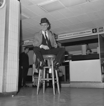 Comedian Dickie Henderson uses a stool as a prop while he waits for his plane at London Airport.